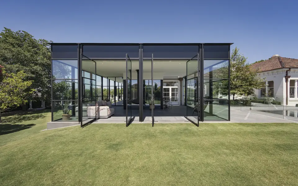 Glasshus concept visual: a black-framed glass pavilion with a thin flat roof on a green lawn under a clear blue sky