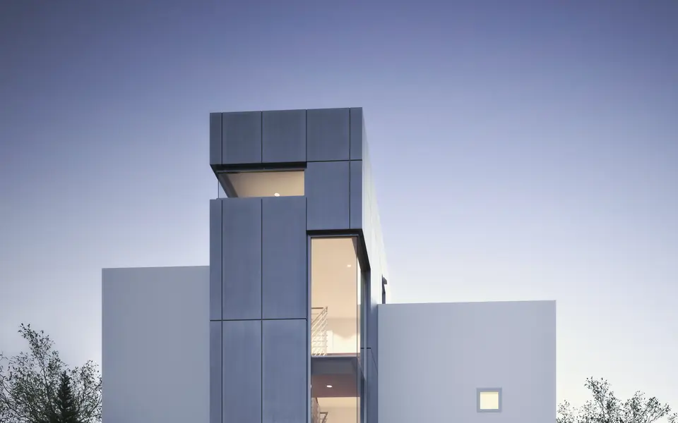 Kube concept visual: a white house of stacked cubic volumes with a lit full-height glazed slot, above a bank of boulders at dusk